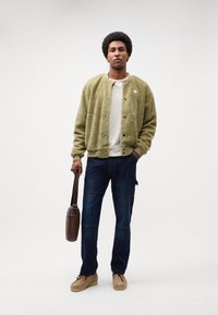 Giacca in pile verde oliva con bottoni, camicia crema, jeans blu scuro e scarpe beige. Borsa a tracolla marrone.