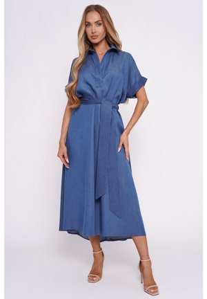 Kobieta stojąca na jednolitym tle, ubrana w niebieską, midi sukienkę z kołnierzykiem i krótkimi rękawami, przewiązaną paskiem, oraz beżowe sandały na wysokim obcasie.