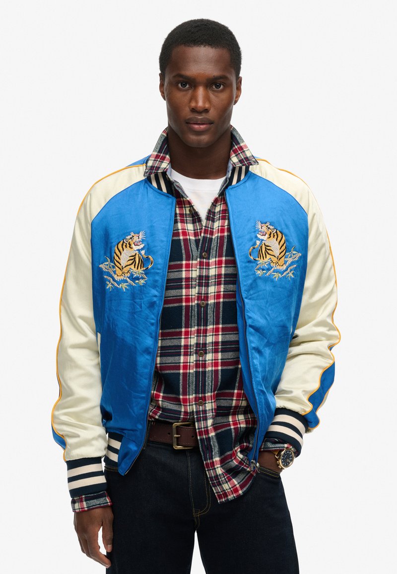 Homme portant une veste bomber bleu et crème avec broderie de tigre sur une chemise à carreaux rouge et marine et un jean noir sur fond blanc.