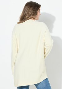 Krémový oversized svetr s ribovanou texturou, dlouhými rukávy a bočními rozparky, v kombinaci s modrými džíny.