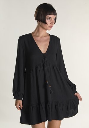 Frau mit kurzen dunklen Haaren, die ein schwarzes, langärmliges Kleid mit tiefem V-Ausschnitt und Quastenbändern trägt und den Saum des Kleides auf einer Seite hält.