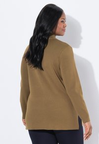 Frau mit langen schwarzen Haaren, die einen braunen Langarmpullover und dunkle Hosen trägt, leicht nach rechts gewandt vor einem weißen Hintergrund.
