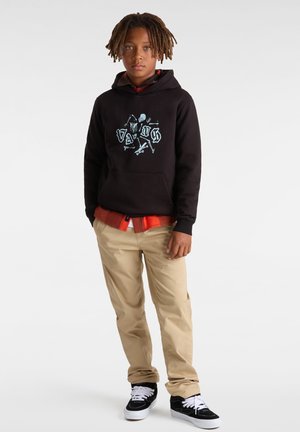 Felpa nera con grafica di scheletro, pantaloni beige e sneakers nere. La felpa ha una tasca frontale ed è sovrapposta a un flanella arancione.