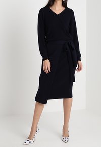 Abito a portafoglio navy realizzato in tessuto morbido, con scollo a V profondo, maniche lunghe, dettaglio vita legato e un orlo asimmetrico alla lunghezza del ginocchio.