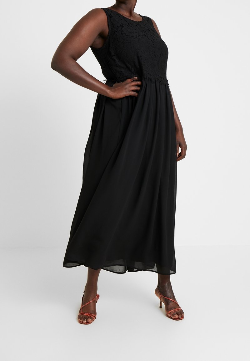 Femme portant une robe noire sans manches en dentelle avec une jupe fluide et des sandales rouges à talons hauts avec des lanières, debout devant un fond blanc.