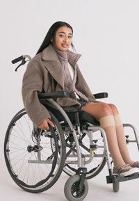 Invalidný vozík so strieborným rámom a veľkými kolesami. Osoba má na sebe hnedý kabát cez prešívanú bundu, doplnené béžovými pančuchami po kolená.