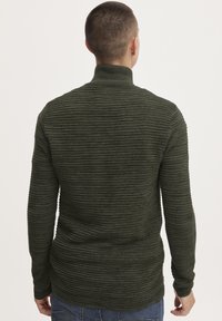 Mørkegrøn ribbet turtleneck sweater med lange ærmer og en struktureret overflade, tætsiddende design og høj krave, parret med blå jeans.