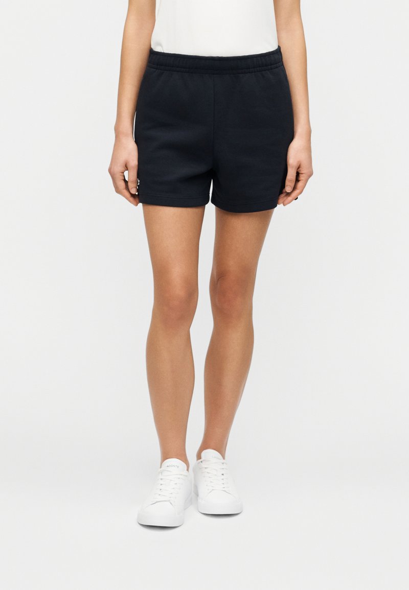 Shorts en coton noirs avec une taille élastique, présentant une texture lisse et des poches latérales, complétés par des baskets blanches.