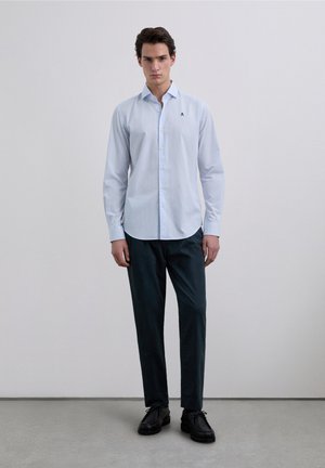 Camicia azzurro chiaro con bottoni, taglio slim e maniche lunghe, con un piccolo logo sul petto a sinistra. Abbinata a pantaloni verde scuro.