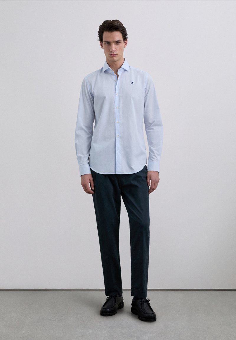 Lichtblauw overhemd met knopen, een slanke pasvorm en lange mouwen, voorzien van een klein logo op de linkerborst. Gecombineerd met donkergroene broek.