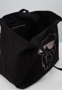 Black canvas tote bag featuring an embroidered graphic of a face wearing sunglasses. Includes a zippered pocket inside for storage.