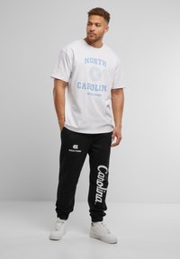 Hvid bomulds t-shirt med blå tekst "NORTH CAROLINA" og et segl. Sorte sweatpants med hvid "Carolina" tryk og logo. Hvide sneakers.