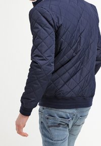 Osoba ubrana w granatową pikowaną kurtkę i jasnoniebieskie dżinsy, pokazana od tyłu z lekkim obrotem w lewo.