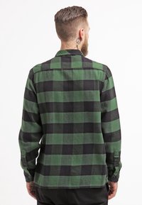 Man med kort hår och skägg, klädd i en grön och svart rutmönstrad långärmad skjorta, vänd bort från kameran mot en enkel vit bakgrund.