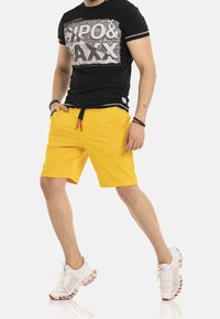 Czarny t-shirt z grafiką, w połączeniu z jasnymi żółtymi szortami z kieszeniami oraz elastycznym sznurkiem. Białe buty sportowe.
