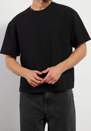 Mężczyzna w gładkiej czarnej koszulce z krótkim rękawem i ciemnych dżinsach, z dłońmi luźno skrzyżowanymi na biodrach, na tle jasnoszarym.