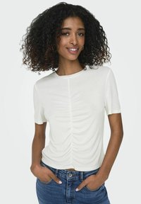 Camiseta blanca de manga corta con detalle fruncido en el centro. Tejido suave, escote redondo y corte relajado. Combinada con jeans de mezclilla azules.