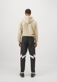 Sudadera con capucha beige combinada con pantalones deportivos negros que presentan rayas diagonales en color burdeos y blanco, tejido ligero y corte ajustado.