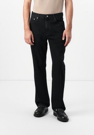 Jeans de mezclilla negra con un corte recto, cinco bolsillos y herrajes metálicos. Combinados con una camisa beige y zapatos negros.