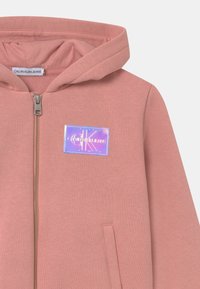 Felpa rosa con zip realizzata in tessuto morbido, dotata di una tasca frontale, un patch logo riflettente e cappuccio regolabile con coulisse.