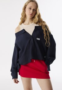 Marineblauwe oversized button-up shirt van een gestructureerde stof, met een off-shoulder ontwerp en een witte logo patch, gecombineerd met een aansluitende rode minirok.