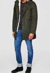 Olivgrön parkasjacka med fleecefoder, knäppning med knappar, huva och sidofickor, matchad med blå jeans och svarta sneakers.