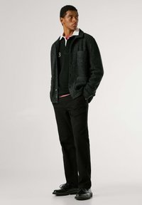 Donkergrijs jack met knopen en kraag, zwarte trui met witte accenten, zwarte broek en zwarte schoenen, die samen een gelaagde, casual look creëren.