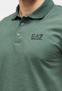 Polo de algodão verde com pala de três botões, logótipo "EA7" bordado e textura suave.