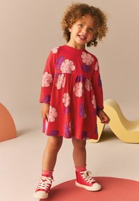 Rochie din bumbac roșu cu mâneci lungi, având modele de nori roz și mov. Poartă adidași roșii și șosete roz până la glezne.