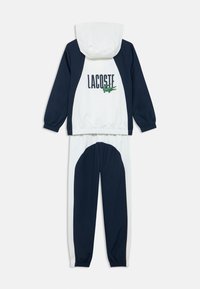 Modro-biely teplákový komplet s mikinou s kapucňou s logom "LACOSTE" a zeleným krokodílím motívom, doplnený o zodpovedajúce nohavice. Elastické manžety.