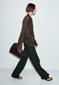 Donna che cammina in una blusa trasparente marrone, pantaloni neri larghi, sandali a infradito, mentre porta una grande borsa a tracolla marrone su uno sfondo neutro.