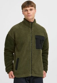 Fleece jacket in olive green with a zip front, high collar, black chest pocket, and side pockets, featuring a textured, soft exterior.