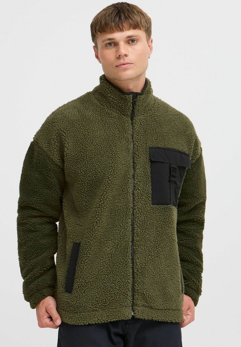 Fleece jacket in olive green with a zip front, high collar, black chest pocket, and side pockets, featuring a textured, soft exterior.