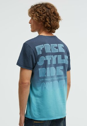 Junger Mann mit lockigem Haar, der ein kurzärmeliges blaues Ombre-T-Shirt mit dem Schriftzug "FREE STYLE RIDE" auf dem Rücken trägt.