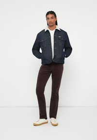 Giubbotto di jeans con un colletto di pelliccia sintetica di colore chiaro, caratterizzato da tasche frontali, abbinato a pantaloni di velluto a coste marrone scuro e sneaker bianche.