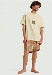 T-shirt de algodão bege com gráfico quadrado em tons terrosos, combinado com calções padrão em flores laranja e verdes sobre um fundo bege.