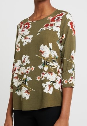 Kobieta w oliwkowozielonym topie z długim rękawem z dużym białym i czerwonym kwiatowym nadrukiem, w czarnych spodniach, stojąca na jednolitym tle.