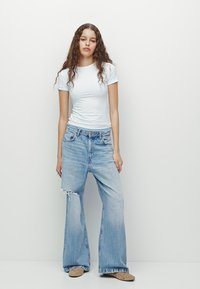 Weißes, eng anliegendes T-Shirt kombiniert mit hellblauen, weit geschnittenen Jeans mit fransigem Saum. Das Model trägt beigen Slipper mit Verzierungen.