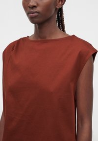 Femme à la peau foncée et aux cheveux tressés portant un haut sans manches couleur rouille sur un fond uni.