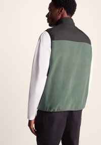 Fleece vest in green with a black upper section, featuring a high collar and contrasting white long sleeves, shown from the back.