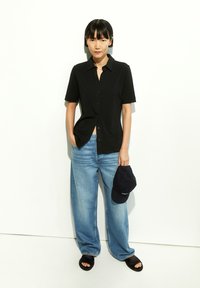 Black short-sleeve button-up shirt, light blue wide-leg jeans, black baseball cap, and black slide sandals against a white background.