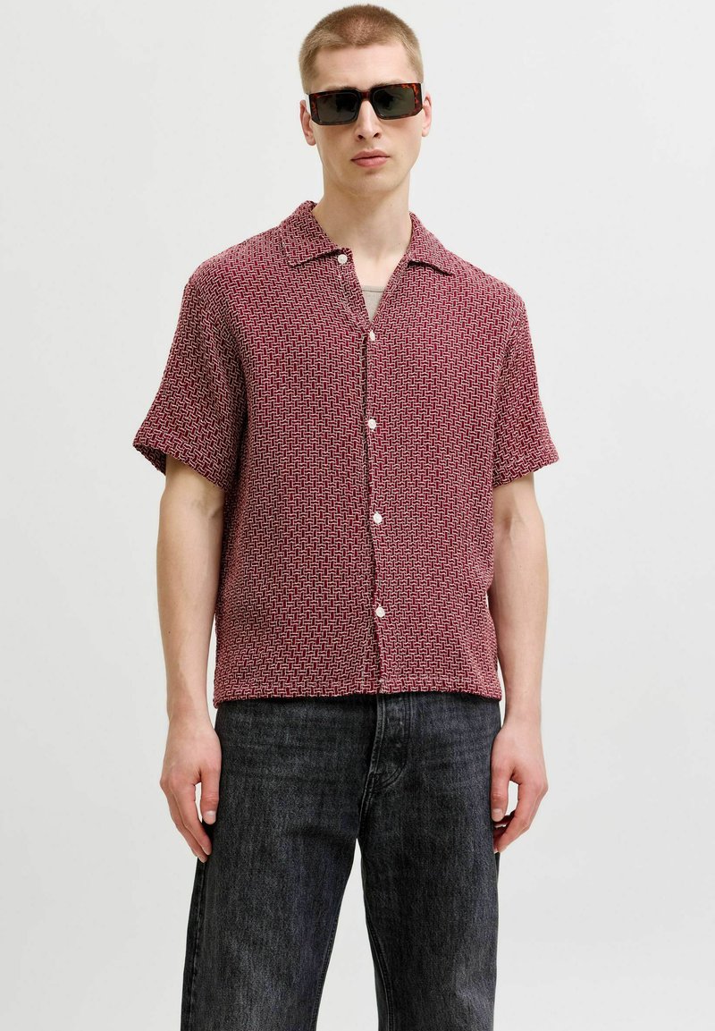 Homme portant une chemise rouge à manches courtes à boutons avec motifs, un jean foncé et des lunettes de soleil, debout devant un fond clair uni.