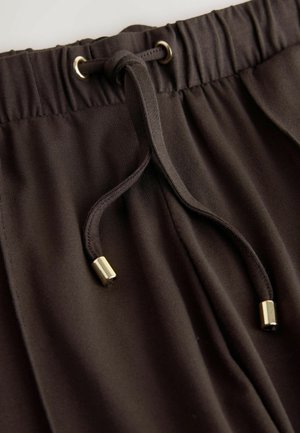 Barna zsinóros nadrág, puha anyagból, sima textúrával, rugalmas derékpánttal és fém díszítésekkel a zsinórvégeken.