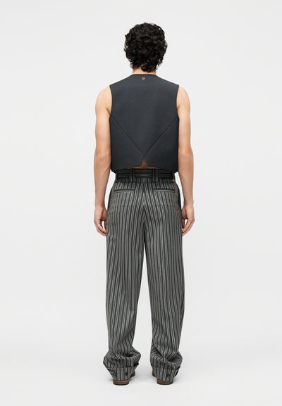 Homme aux cheveux bouclés vu de dos, portant un gilet noir sans manches et un pantalon gris ample à fines rayures avec des poches arrière.