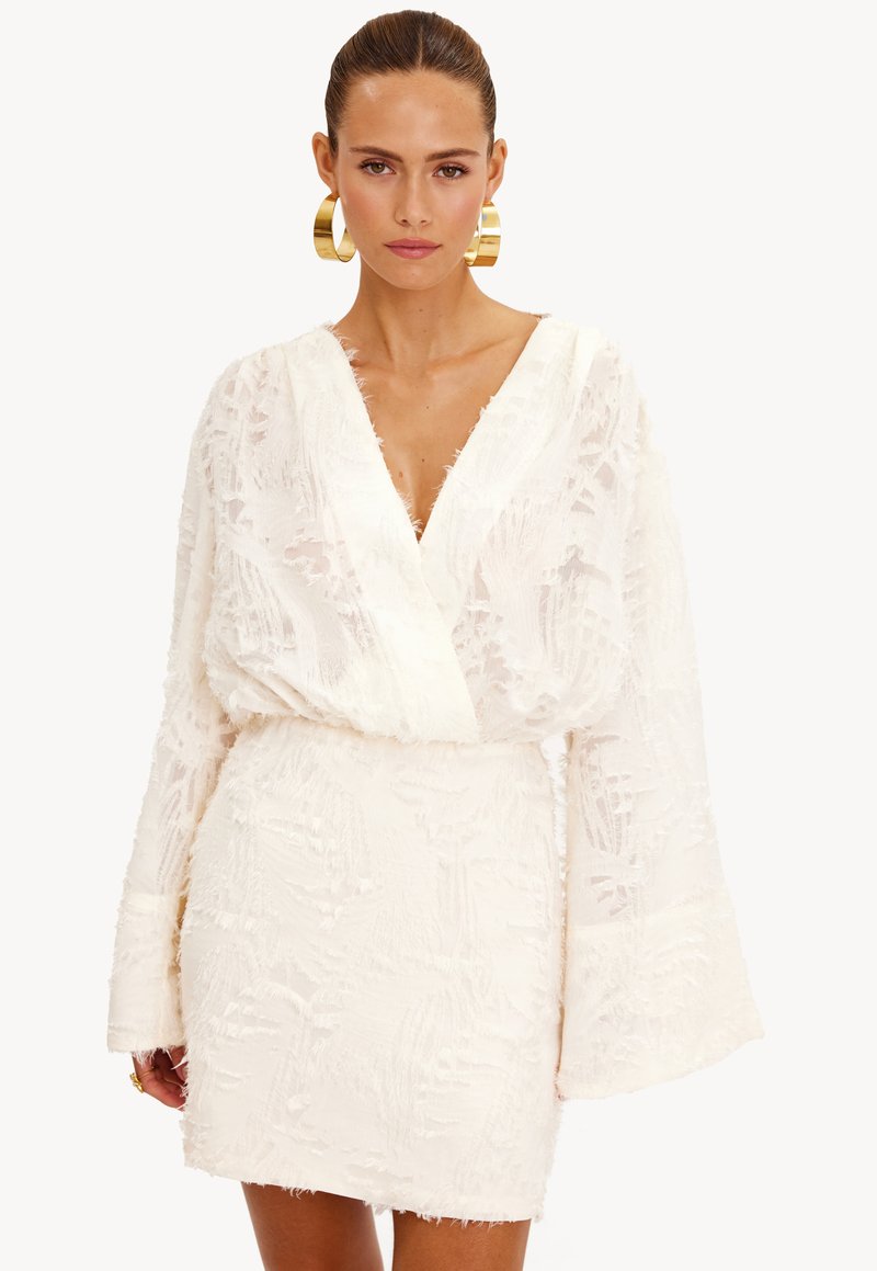 Witte, gestructureerde mini-jurk met een wikkelhalslijn en lange, brede mouwen. Bevat franjes en een taillering. Gouden oorringen.
