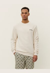 Felpa crema con maniche lunghe e colletto rotondo, caratterizzata da un piccolo logo ricamato sul petto. Abbinata a pantaloni corti a fantasia.
