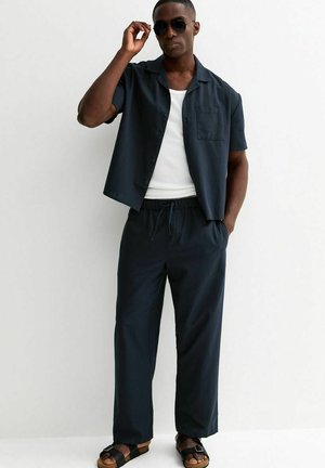 Mann trägt schwarze Sonnenbrille, schwarzes kurzärmeliges Hemd mit Knöpfen über weißem Tanktop, schwarze Kordelzughose und schwarze Sandalen.