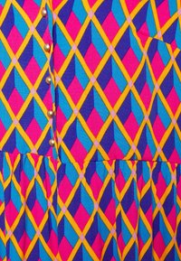 Kolorowa tkanina sukienkowa z geometrycznym wzorem diamentów w niebieskim, różowym, purpurowym i żółtym, z ozdobnymi guzikami wzdłuż szwu.