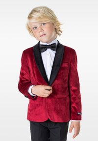 Chaqueta de esmoquin de terciopelo rojo con solapa negra, cierre de un solo botón y detalles en los bolsillos. Llevada sobre una camisa blanca y una pajarita negra.