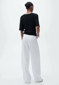 Vrouw met rug naar de camera, gekleed in een zwart is kort mouw gebreid top en witte wijde broek, met handen in de zakken, neutrale achtergrond.